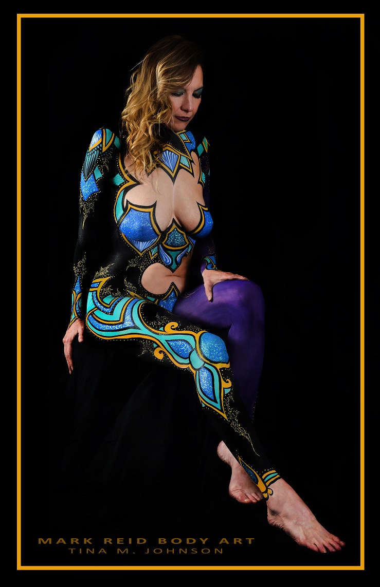 Beautiful long haired sandy blonde woman with a brightly colored full body painting that is black blue tuquoise and gold with lots of gold detailing